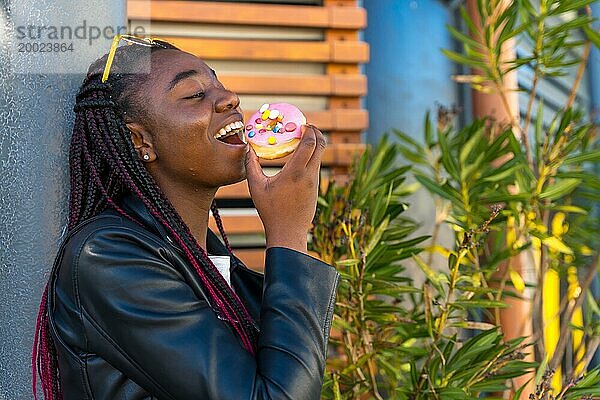 Fröhliche afrikanische Frau mit Lederjacke  die an einer Straßenecke einen Donut isst