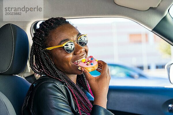 Bezaubernde afrikanische Frau lächelt  während sie in einem Auto sitzend einen Doughnut isst