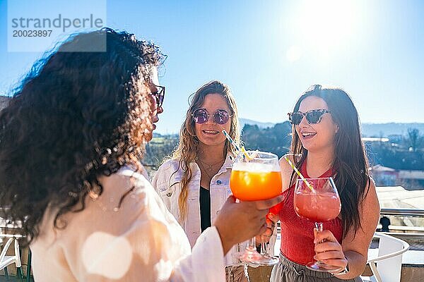 Drei coole Freundinnen stoßen an einem sonnigen Tag mit Cocktails auf einer Dachterrasse auf den Sommer an
