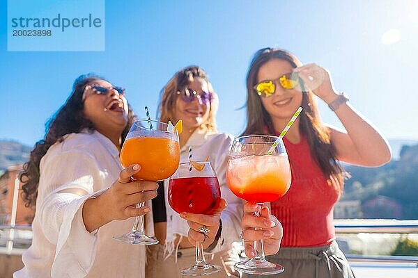 Frontalfoto von drei Freundinnen  die Cocktails trinken und sich auf einem Dach im Sommer amüsieren