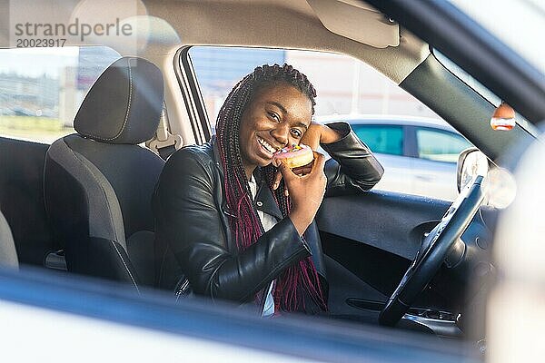 Blick von außen aus dem Fenster einer lächelnden süßen afrikanischen Frau  die in einem Auto Gebäck isst