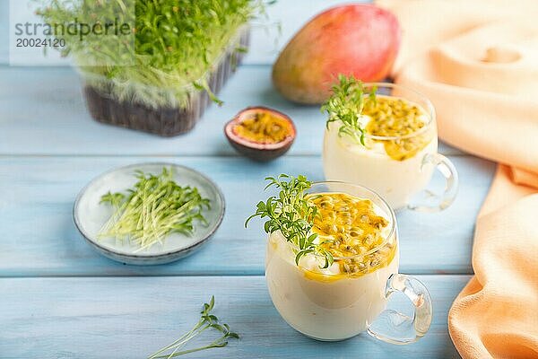 Mango Joghurt mit Passionsfrucht und Koriander Mikrogrün im Glas auf blauem Holzhintergrund mit orangefarbenem Leinenstoff. Seitenansicht  Nahaufnahme  selektiver Fokus