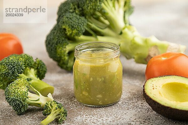 Baby Püree mit Gemüsemischung  Brokkoli  Tomaten  Gurken  Avocado Säuglingsnahrung in Glasgefäß auf braunem Beton Hintergrund. Seitenansicht  Nahaufnahme  selektiver Fokus  Konzept der künstlichen Ernährung