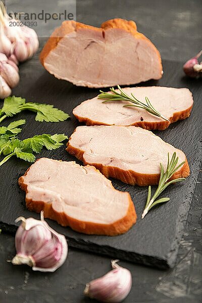 Geräucherter Schweineschinken auf einem Schieferbrett mit Pfeffer  Knoblauch und Kräutern auf schwarzem Betonhintergrund. Seitenansicht  Nahaufnahme  selektiver Fokus