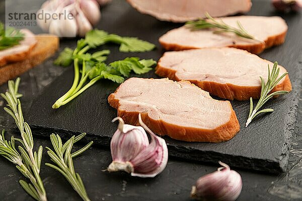 Geräucherter Schweineschinken auf einem Schieferbrett mit Pfeffer  Knoblauch und Kräutern auf schwarzem Betonhintergrund. Seitenansicht  Nahaufnahme  selektiver Fokus