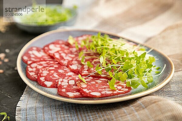 Scheiben von geräucherter Cervelatwurst mit Spinat  Salz und Pfeffer auf schwarzem Betonhintergrund und beigem Textil. Seitenansicht  Nahaufnahme  selektiver Fokus