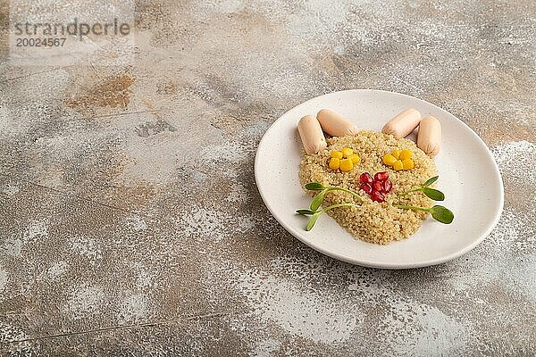 Lustig gemischter Quinoa Brei  Mais  Granatapfelkerne und kleine Würstchen in Form eines Katzengesichts auf braunem Betonhintergrund. Seitenansicht  Kopie Raum. Essen für Kinder  gesundes Essen Konzept