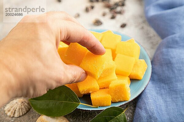 Getrocknete und kandierte Mangowürfel auf blauem Teller mit Hand auf braunem Betonhintergrund und Leinentextil. Seitenansicht  Nahaufnahme  selektiver Fokus  veganes  vegetarisches Lebensmittelkonzept