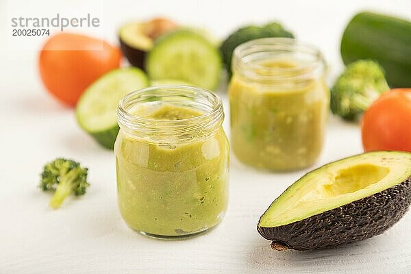 Baby Püree mit Gemüsemischung  Brokkoli  Tomaten  Gurken  Avocado Säuglingsnahrung in Glasgefäß auf weißem Holzhintergrund. Seitenansicht  Nahaufnahme  selektiver Fokus  Konzept der künstlichen Ernährung