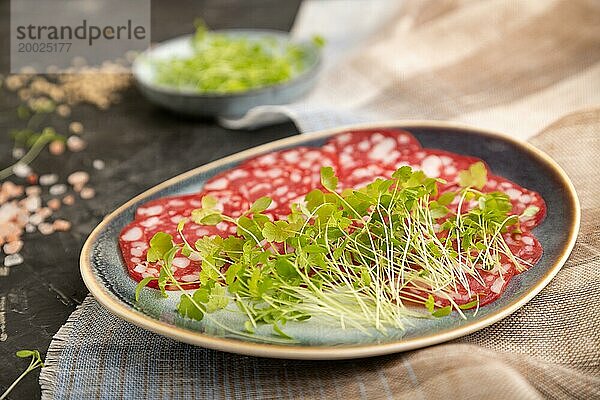 Scheiben von geräucherter Cervelatwurst mit Spinat  Salz und Pfeffer auf schwarzem Betonhintergrund und beigem Textil. Seitenansicht  Nahaufnahme  selektiver Fokus