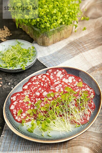 Scheiben von geräucherter Cervelatwurst mit Spinat  Salz und Pfeffer auf schwarzem Betonhintergrund und beigem Textil. Seitenansicht  Nahaufnahme  selektiver Fokus