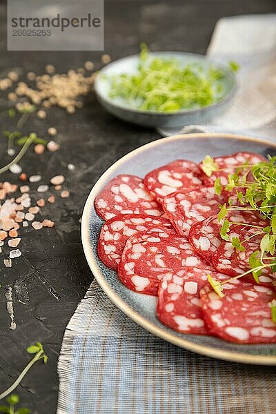 Scheiben von geräucherter Cervelatwurst mit Spinat  Salz und Pfeffer auf schwarzem Betonhintergrund und beigem Textil. Seitenansicht  Nahaufnahme  selektiver Fokus