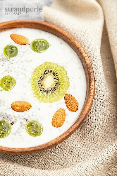 Joghurt mit Kiwi  Stachelbeere  Chia und Mandeln in einer Holzschale auf grauem Holzhintergrund und Leinenstoff. Draufsicht  Nahaufnahme  selektiver Fokus