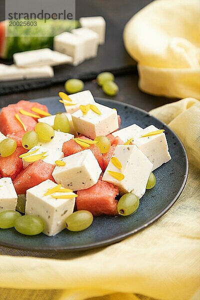 Vegetarischer Salat mit Wassermelone  Fetakäse und Trauben auf blauem Keramikteller auf schwarzem Betonhintergrund und gelbem Leinentuch. Seitenansicht  Nahaufnahme  selektiver Fokus