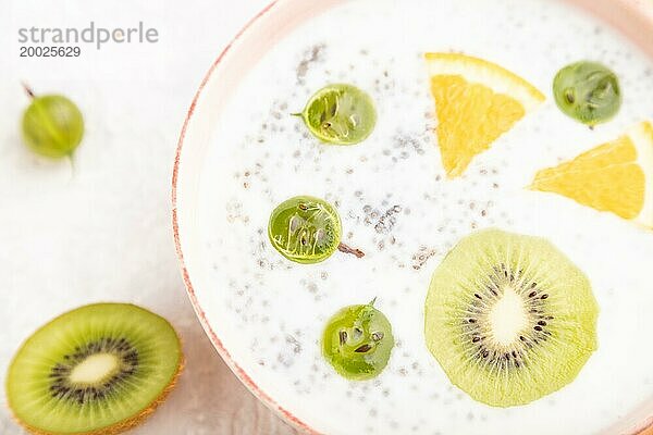 Joghurt mit Kiwi  Stachelbeere und Chia in einer Keramikschale auf grauem Betonhintergrund und orangefarbenem Leinentextil. Draufsicht  Nahaufnahme  selektiver Fokus