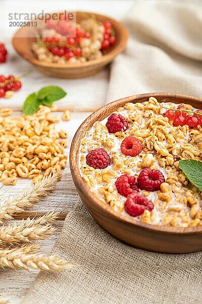 Weizenflockenbrei mit Milch  Himbeeren und Johannisbeeren in einer Holzschale auf weißem Holzhintergrund und Leinentextil. Seitenansicht  Nahaufnahme  selektiver Fokus