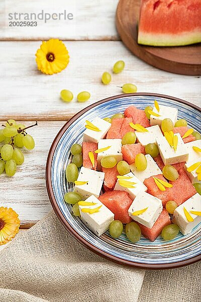 Vegetarischer Salat mit Wassermelone  Fetakäse und Trauben auf blauem Keramikteller auf weißem Holzhintergrund und Leinentuch. Seitenansicht  Nahaufnahme  selektiver Fokus