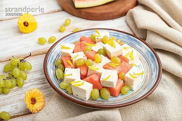 Vegetarischer Salat mit Wassermelone  Fetakäse und Trauben auf blauem Keramikteller auf weißem Holzhintergrund und Leinentuch. Seitenansicht  Nahaufnahme  selektiver Fokus