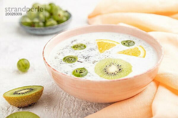 Joghurt mit Kiwi  Stachelbeere und Chia in einer Keramikschale auf grauem Betonhintergrund und orangefarbenem Leinentextil. Seitenansicht  Nahaufnahme  selektiver Fokus