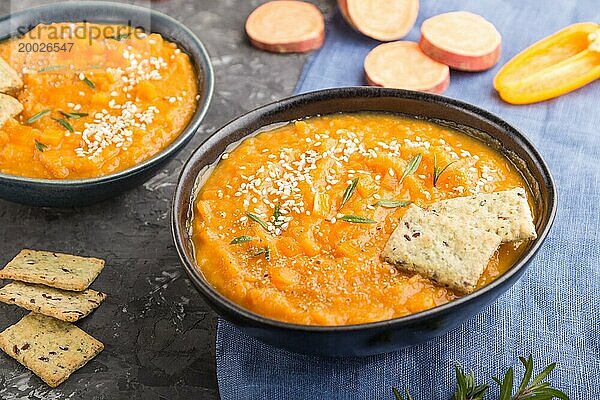 Süßkartoffel oder Batatencremesuppe mit Sesam und Knabbereien in blaün Keramikschalen auf einem schwarzen Betonhintergrund mit blauem Leinentextil. Seitenansicht  Nahaufnahme  selektiver Fokus