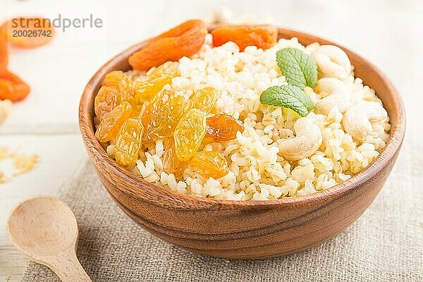 Bulgur Brei mit getrockneten Aprikosen  Rosinen und Cashew in einer Holzschale auf weißem Holzhintergrund und Leinenstoff. Seitenansicht  Nahaufnahme  selektiver Fokus. Traditionelle türkische Küche