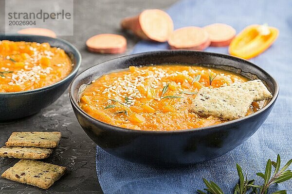 Süßkartoffel oder Batatencremesuppe mit Sesam und Knabbereien in blaün Keramikschalen auf einem schwarzen Betonhintergrund mit blauem Leinentextil. Seitenansicht  Nahaufnahme  selektiver Fokus