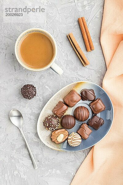 Verschiedene Schokoladenbonbons und eine Tasse Kaffee auf einem grauen Betonhintergrund und orangefarbenem Textil. Draufsicht  Flachlegung  Nahaufnahme