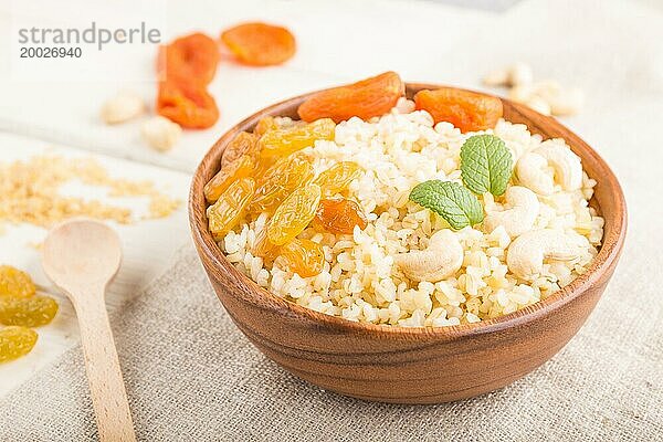 Bulgur Brei mit getrockneten Aprikosen  Rosinen und Cashew in einer Holzschale auf weißem Holzhintergrund und Leinenstoff. Seitenansicht  Nahaufnahme  selektiver Fokus. Traditionelle türkische Küche