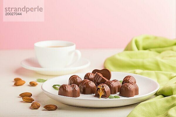 Schokoladenkaramellbonbons mit Mandeln und einer Tasse Kaffee auf weißem und rosa Hintergrund und grünem Textil. Seitenansicht  Kopierraum