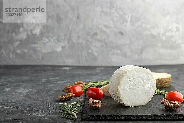 Weißer Ziegenkäse und verschiedene Käsesorten mit Rosmarin und Tomaten auf einer schwarzen Schiefertafel auf einem schwarzen Betonhintergrund. Seitenansicht  Kopierraum  selektiver Fokus