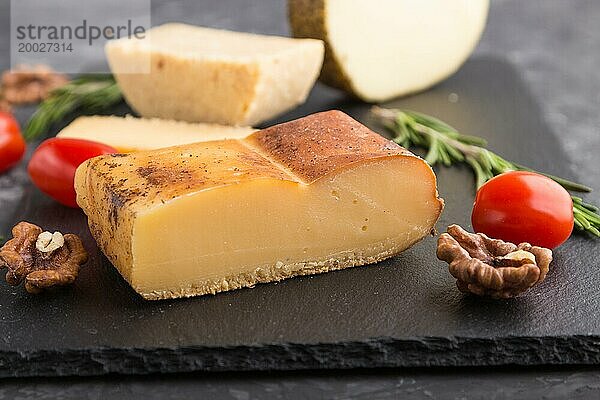 Geräucherter Käse und verschiedene Käsesorten mit Rosmarin und Tomaten auf einer schwarzen Schieferplatte auf einem schwarzen Betonhintergrund. Seitenansicht  Nahaufnahme  selektiver Fokus