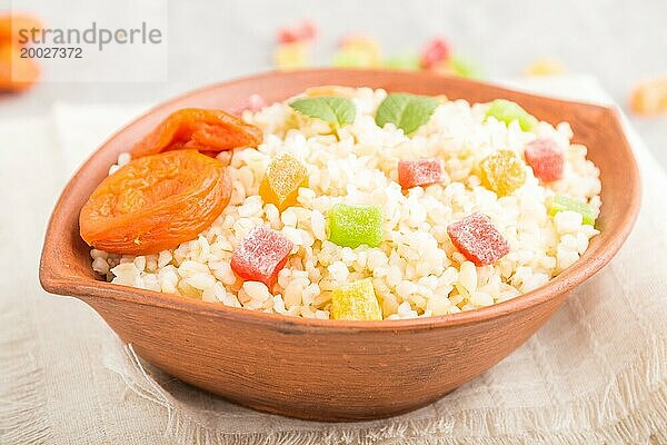 Bulgur Brei mit getrockneten Aprikosen und kandierten Früchten in einer Tonschüssel auf grauem Betonhintergrund und Leinentextil. Seitenansicht  Nahaufnahme  selektiver Fokus. Traditionelle türkische Küche