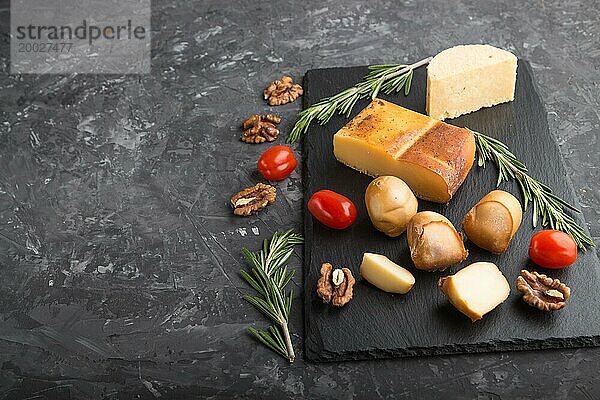 Geräucherter Käse und verschiedene Käsesorten mit Rosmarin und Tomaten auf einer schwarzen Schiefertafel auf einem schwarzen Betonhintergrund. Seitenansicht  Kopierraum  selektiver Fokus