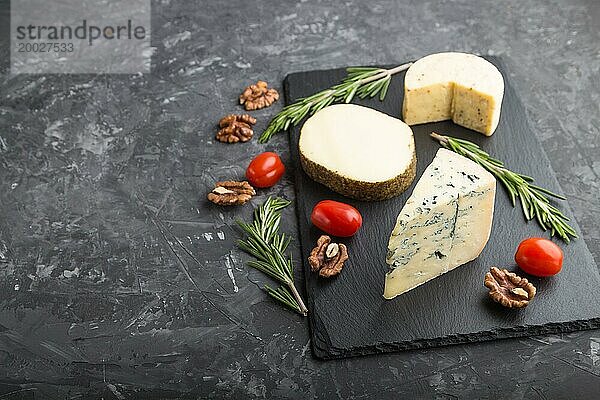 Blauschimmelkäse und verschiedene Käsesorten mit Rosmarin und Tomaten auf einer schwarzen Schiefertafel auf einem schwarzen Betonhintergrund. Seitenansicht  Kopierraum  selektiver Fokus