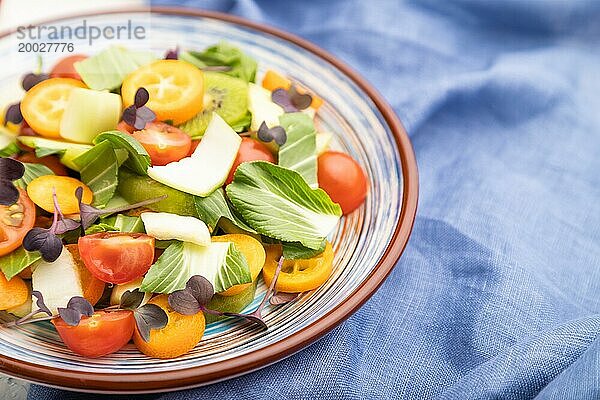 Vegetarischer Salat aus Pac Choi Kohl  Kiwi  Tomaten  Kumquat  Mikrogrünsprossen auf weißem Betonhintergrund und blauem Leinentuch. Seitenansicht  Nahaufnahme  selektiver Fokus