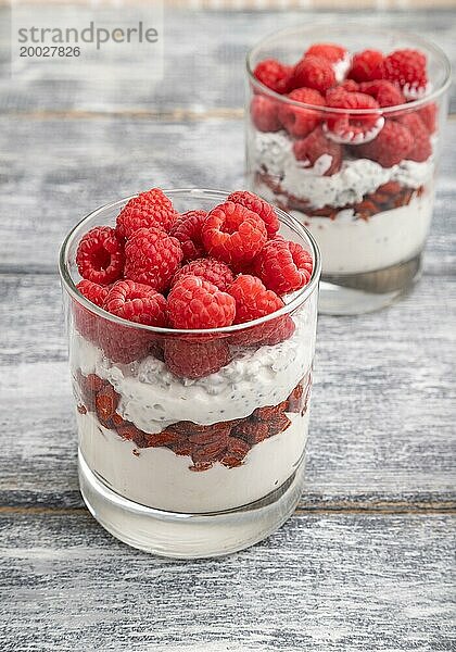 Joghurt mit Himbeeren  Goji Beeren und Chia Samen im Glas auf grauem Holzhintergrund. Seitenansicht  Nahaufnahme  selektiver Fokus