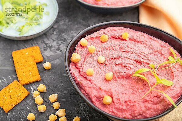 Hummus mit Rüben und mikrogrünen Basilikumsprossen in einer Keramikschale auf einem schwarzen Betonhintergrund und orangefarbenem Textil. Seitenansicht  Nahaufnahme  selektiver Fokus