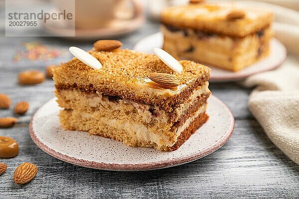 Honigkuchen mit Milchcreme  Karamell  Mandeln und einer Tasse Kaffee auf einem grauen Holzhintergrund und Leinenstoff. Seitenansicht  Nahaufnahme  selektiver Fokus