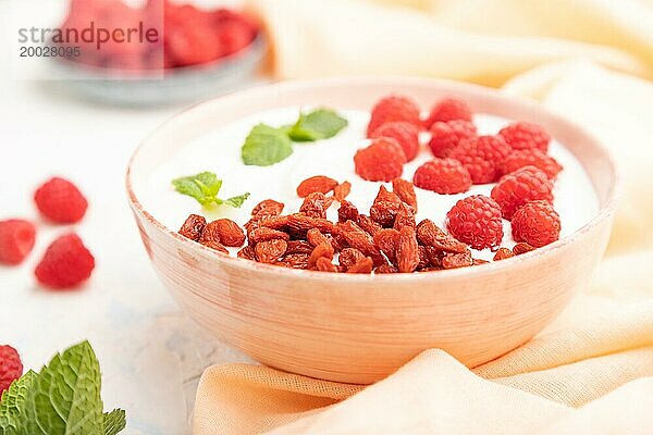 Joghurt mit Himbeeren und Goji Beeren in einer Keramikschale auf weißem Betonhintergrund und orangefarbenem Leinentextil. Seitenansicht  Nahaufnahme  selektiver Fokus