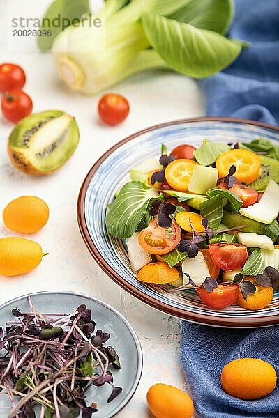 Vegetarischer Salat aus Pac Choi Kohl  Kiwi  Tomaten  Kumquat  Mikrogrünsprossen auf weißem Betonhintergrund und blauem Leinentuch. Seitenansicht  Nahaufnahme  selektiver Fokus