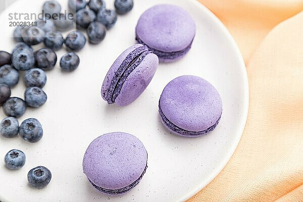 Lila Macarons oder Makronen Kuchen mit Blaubeeren auf weißem Keramikteller auf einem grauen Beton Hintergrund und orange Textil. Draufsicht  Nahaufnahme  selektiver Fokus