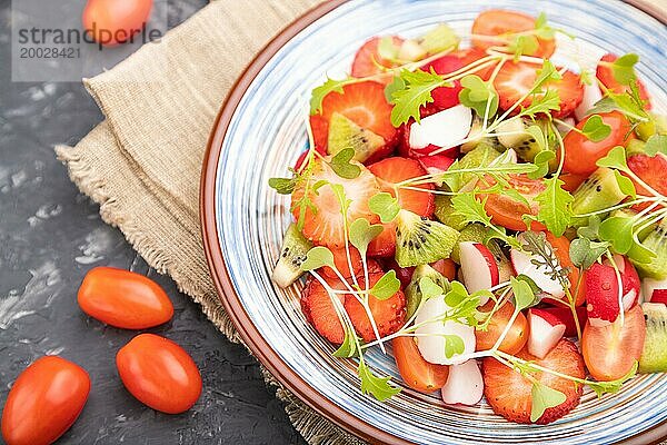 Vegetarischer Obst und Gemüsesalat aus Erdbeeren  Kiwi  Tomaten  Mikrogrünsprossen auf schwarzem Betonhintergrund und Leinentextil. Seitenansicht  Nahaufnahme  selektiver Fokus