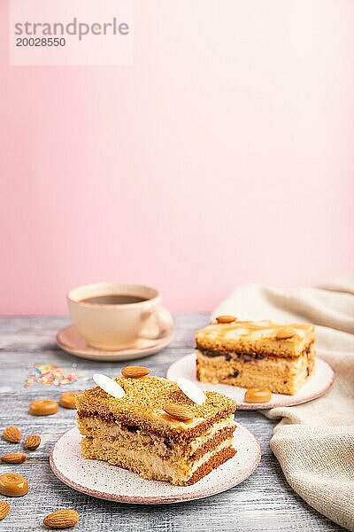 Honigkuchen mit Milchcreme  Karamell  Mandeln und einer Tasse Kaffee auf grauem und rosa Hintergrund und Leinenstoff. Seitenansicht  Nahaufnahme  selektiver Fokus  Kopierraum