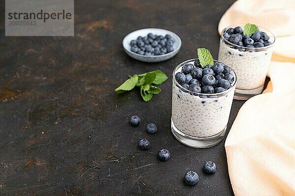 Joghurt mit Heidelbeeren und Chia im Glas auf schwarzem Betonhintergrund und orangefarbenem Leinentextil. Seitenansicht  Kopierraum