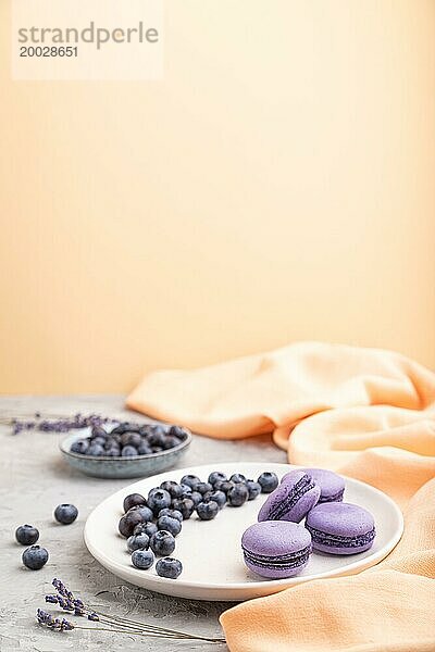 Lila Macarons oder Makronen Kuchen mit Blaubeeren auf weißem Keramikteller auf einem grauen und orangefarbenen Hintergrund und orangefarbenen Textil. Seitenansicht  Kopierraum  selektiver Fokus