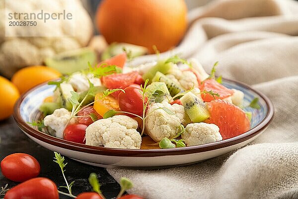 Vegetarischer Salat aus Blumenkohl  Kiwi  Tomaten  Mikrogrünsprossen auf schwarzem Betonhintergrund und Leinentuch. Seitenansicht  Nahaufnahme  selektiver Fokus