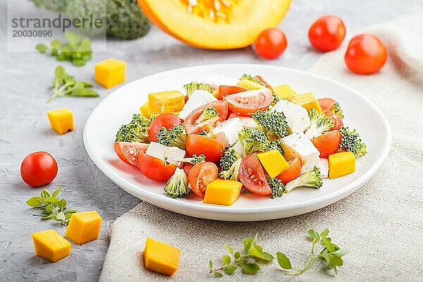 Vegetarischer Salat mit Brokkoli  Tomaten  Fetakäse und Kürbis auf weißem Keramikteller auf grauem Betonhintergrund und Leinentuch  Seitenansicht  Nahaufnahme  selektiver Fokus