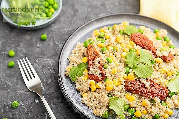 Quinoa Brei mit grünen Erbsen  Mais und getrockneten Tomaten auf einem Keramikteller vor einem grauen Betonhintergrund und gelbem Textil. Seitenansicht  Nahaufnahme  selektiver Fokus