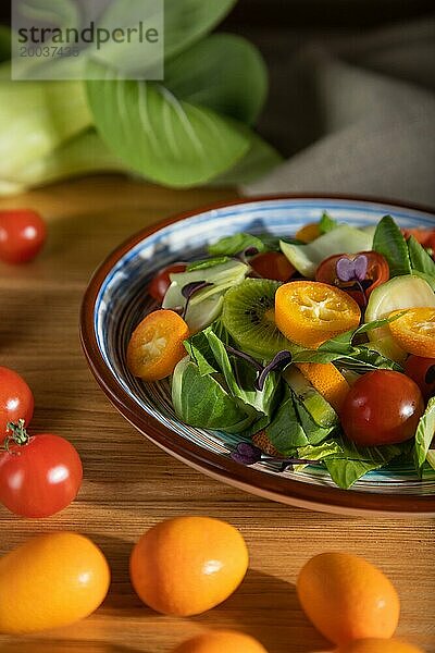 Vegetarischer Salat aus Pac Choi Kohl  Kiwi  Tomaten  Kumquat  mikrogrünen Sprossen auf einem hölzernen Hintergrund und Leinenstoff. Hartes Licht  Kontrast. Seitenansicht  Nahaufnahme  selektiver Fokus