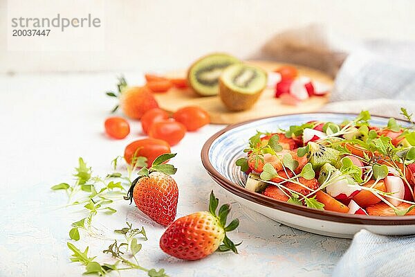 Vegetarischer Obst und Gemüsesalat aus Erdbeeren  Kiwi  Tomaten  Mikrogrünsprossen auf weißem Betonhintergrund und Leinentextil. Seitenansicht  Nahaufnahme  selektiver Fokus